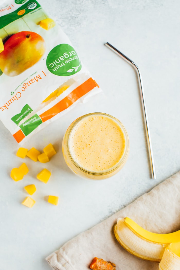 Bright yellow smoothie in a mason jar with a stainless steel straw. In the background there are frozen mango chunks, a banana, fresh ginger and turmeric.