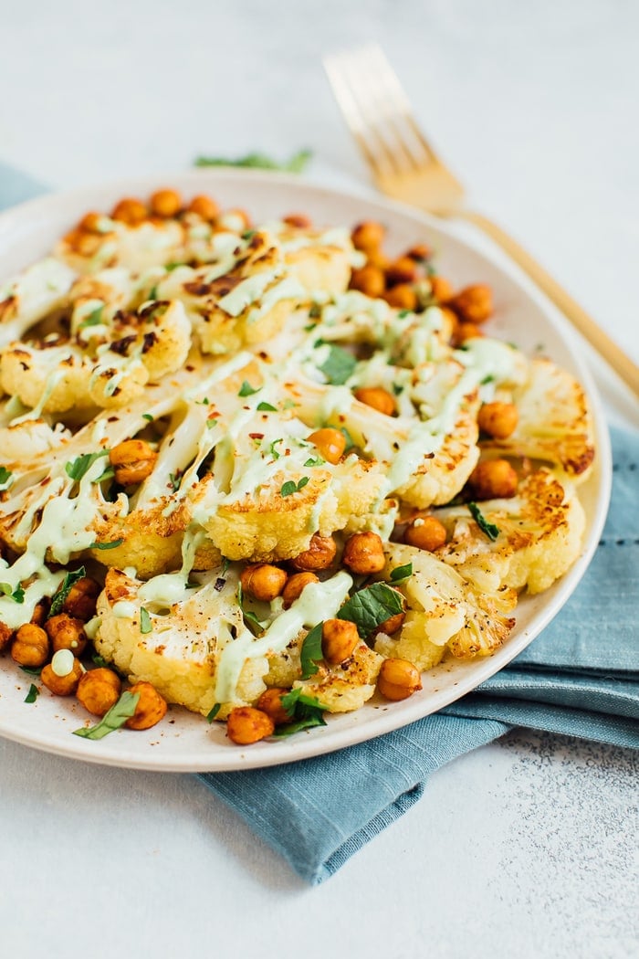 Roasted cauliflower steaks on a tan plate with roasted chickpeas and a green tahini sauce.