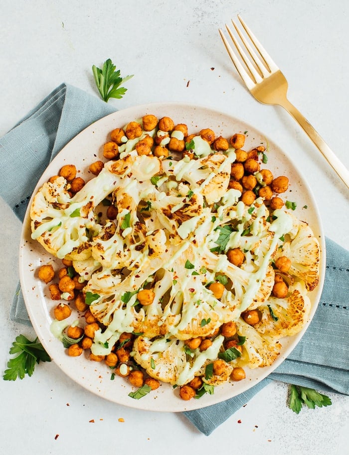 Roasted cauliflower steaks on a tan plate with roasted chickpeas and a green tahini sauce.
