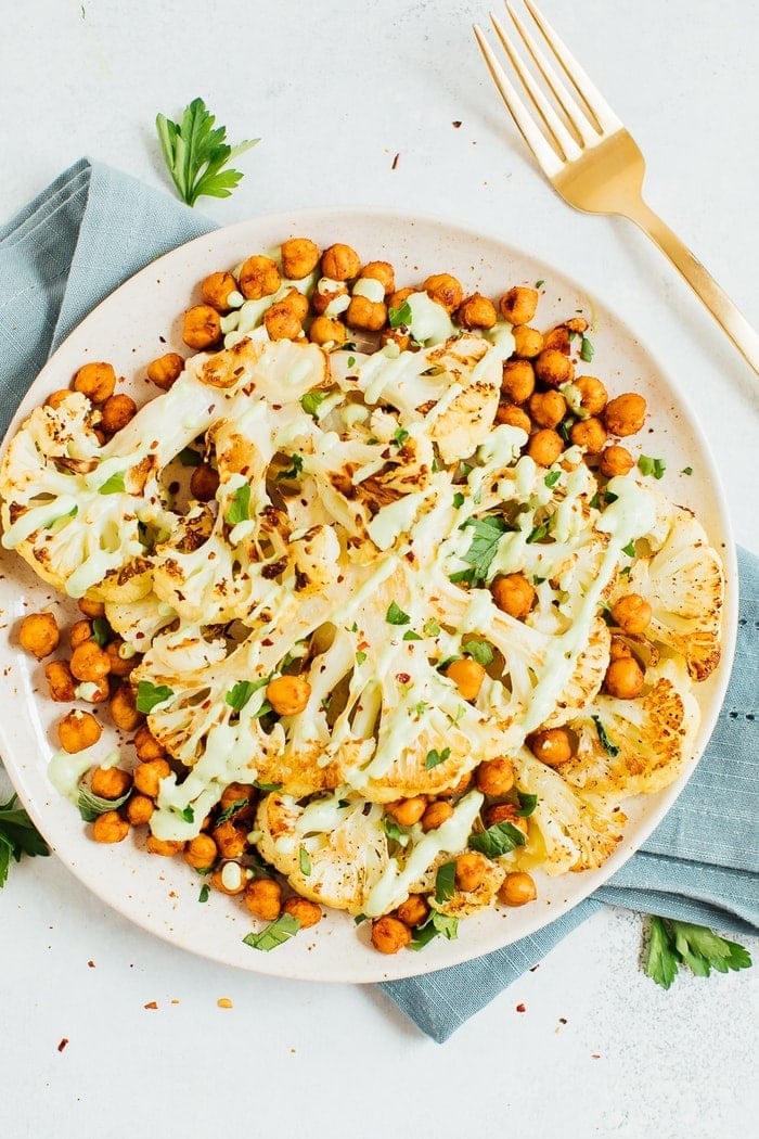 Cauliflower steaks with roasted chickpeas and a green tahini sauce on a plate with a gold fork in the background. 