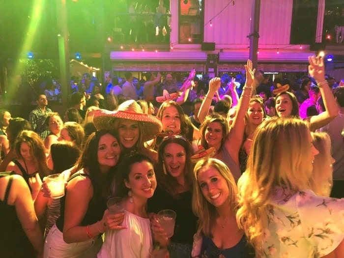 A shot of 10 women in a crowded bar, smiling. Some are wearing sombreros. 