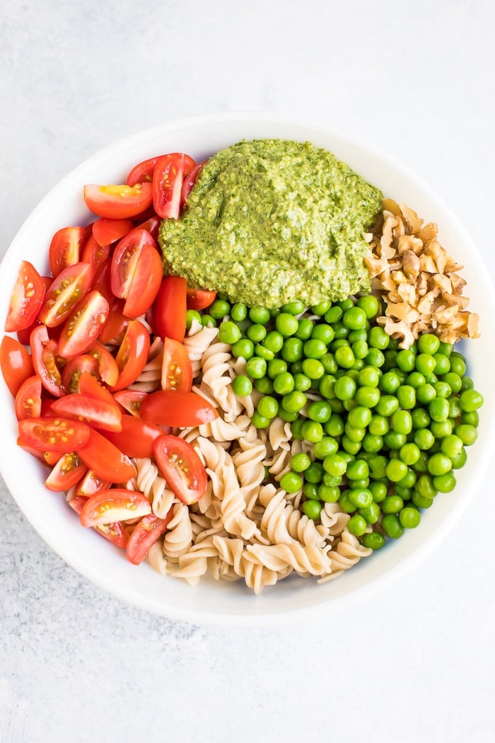 Pesto pasta with tomatoes, peas, walnuts, and pesto separated in a white bowl.
