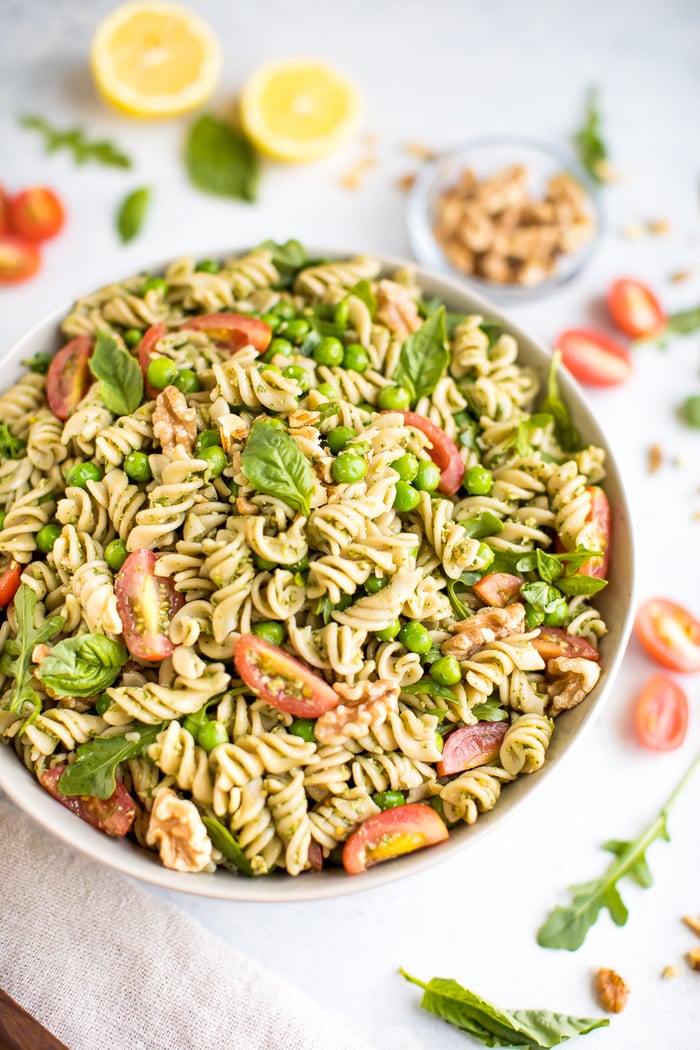 Pesto pasta with tomatoes, peas, and basil in a white bowl, surrounded by lemon, tomatoes, and a bowl of walnuts.