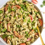 Pesto pasta with tomatoes, peas, and basil in a white bowl, surrounded by tomatoes and arugala.