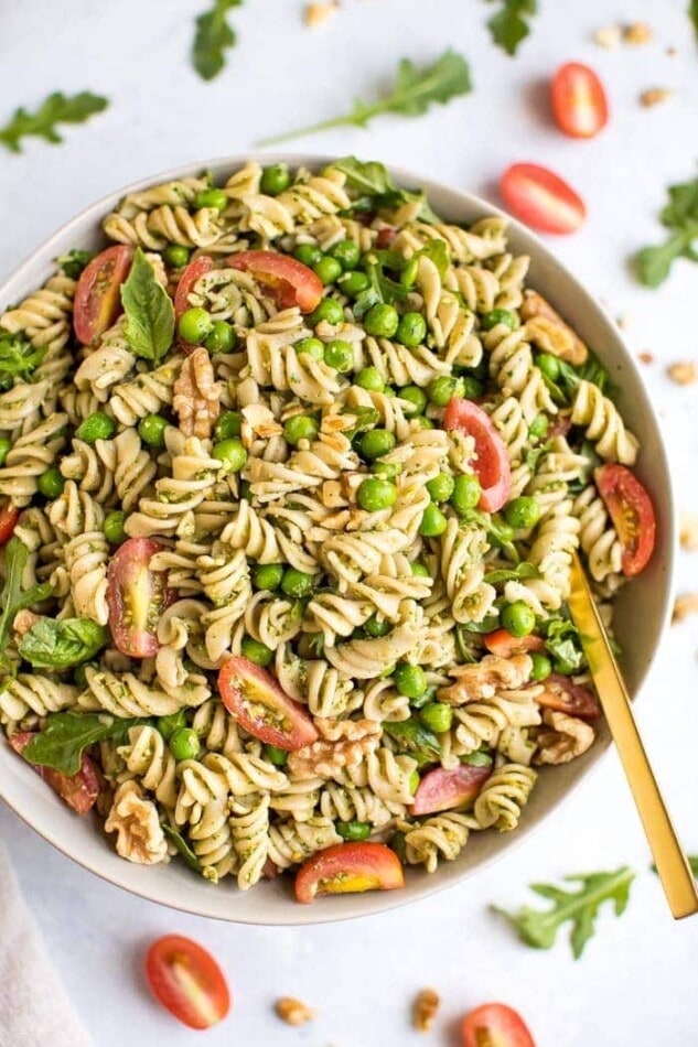 Pesto pasta with tomatoes, peas, and basil in a white bowl, surrounded by tomatoes and arugala.