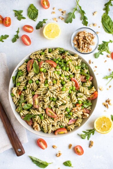 Pesto pasta with tomatoes, peas, and basil in a white bowl, surrounded by lemon, tomatoes, and a bowl of walnuts.