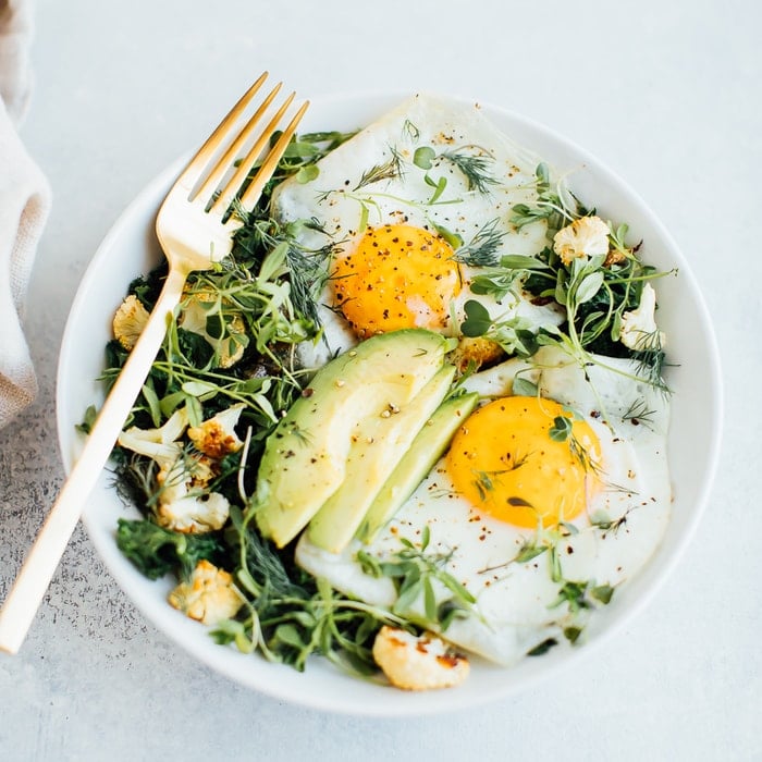 A white bowl with sautéed greens, roasted cauliflower, two sunny side up eggs, avocado, microgreens and fresh dill. 