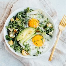 Greens topped with over easy eggs, avocado and roasted cauliflower in a white bowl with a fork on a neutral napkin.