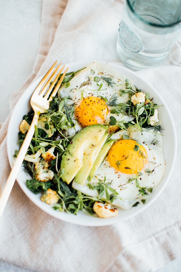 A white bowl with sautéed greens, roasted cauliflower, two sunny side up eggs, avocado, microgreens and fresh dill. 