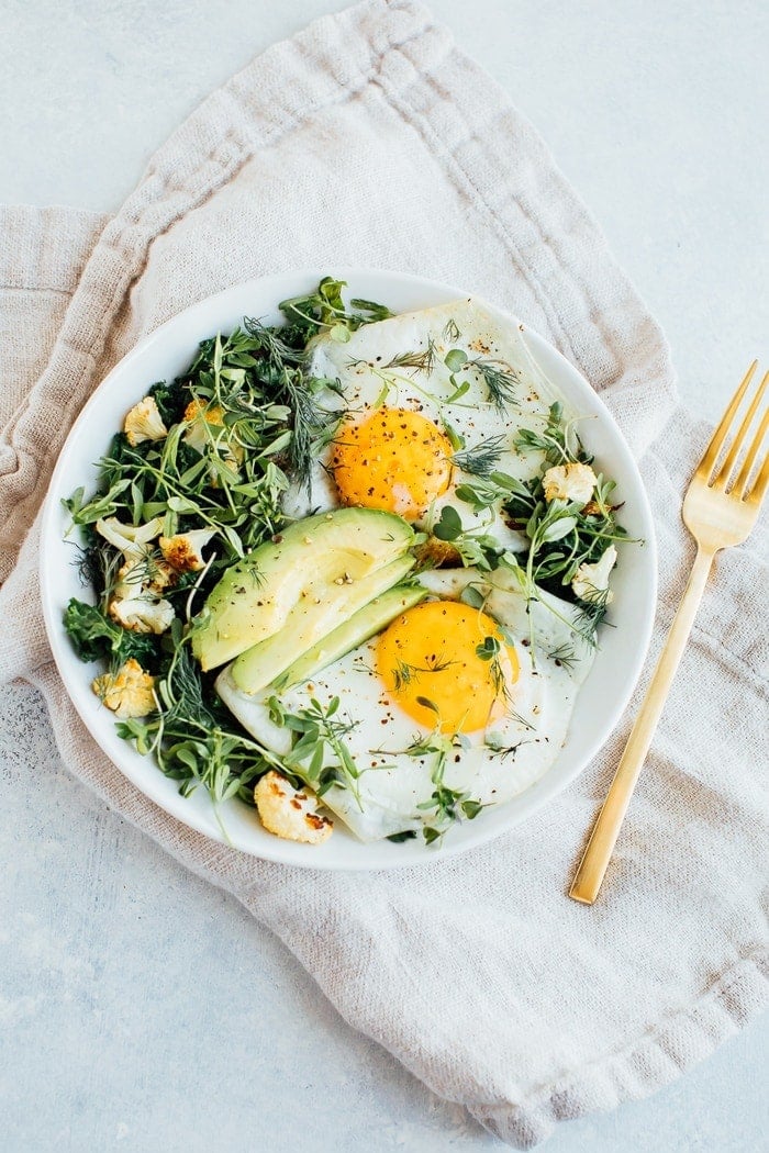 A white bowl with sautéed greens, roasted cauliflower, two sunny side up eggs, avocado, microgreens and fresh dill. 