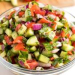 Cucumber, tomato, red onion salad in a glass bowl, next to a cutting board with chopped cucumber and tomato.