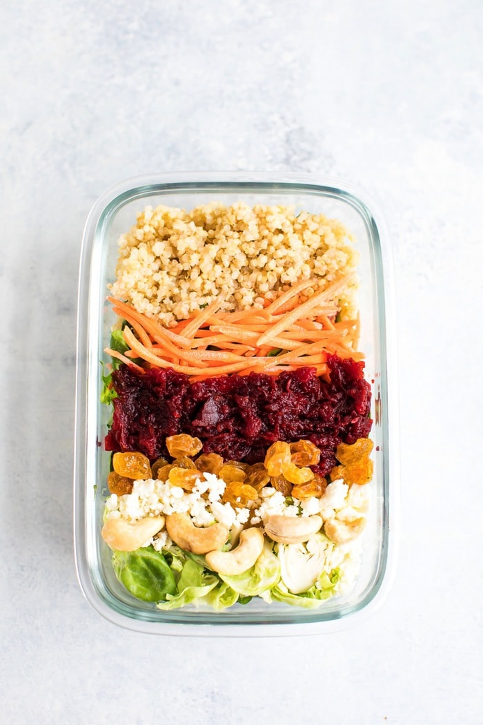 Beet power salad in a meal prep container with quinoa, carrots, beets, raisins, goat cheese, cashews and shredded sprouts on top.