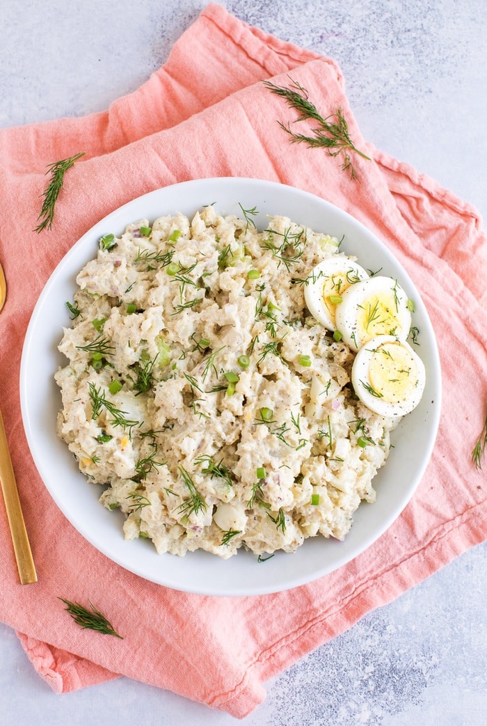 Cauliflower potato salad in a white bowl with three slices of hardboiled egg on the side. 