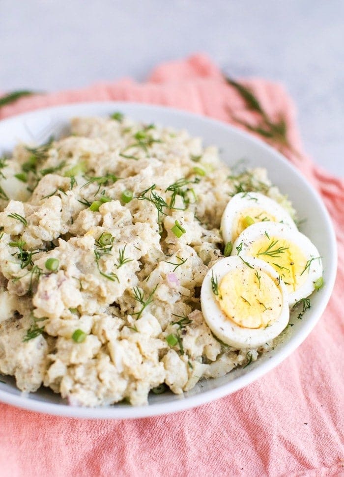 Cauliflower potato salad in a white bowl with three slices of hardboiled egg on the side. 
