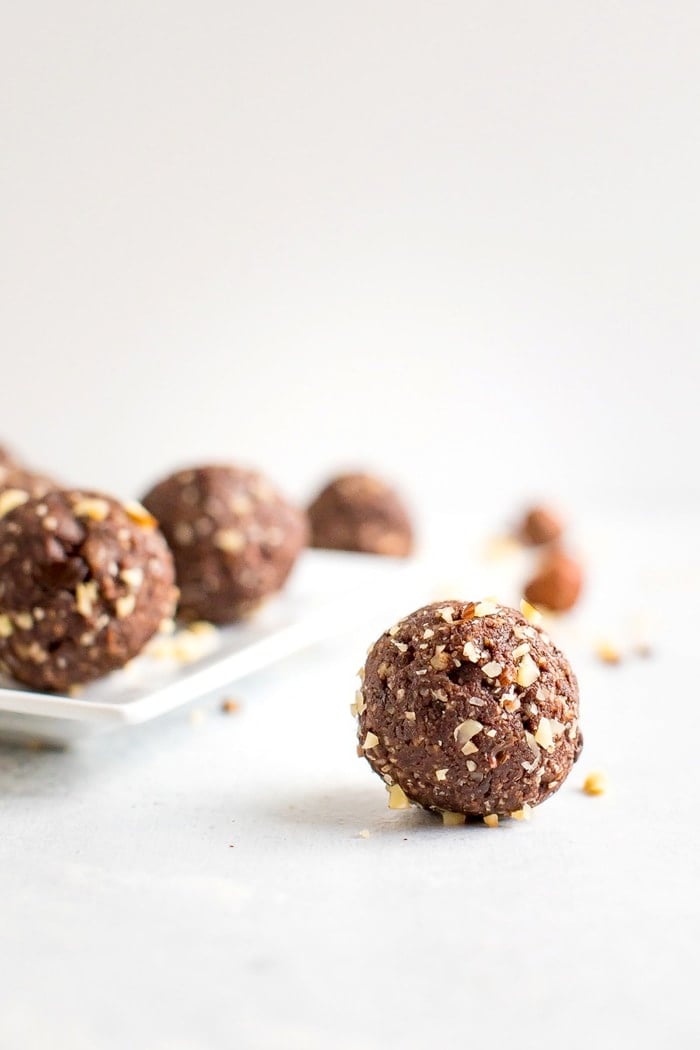 PIN THIS --> Healthy Homemade Ferrero Rocher Bites Homemade chocolate hazelnut energy bite in front with additional energy bites in the background on a white plate.