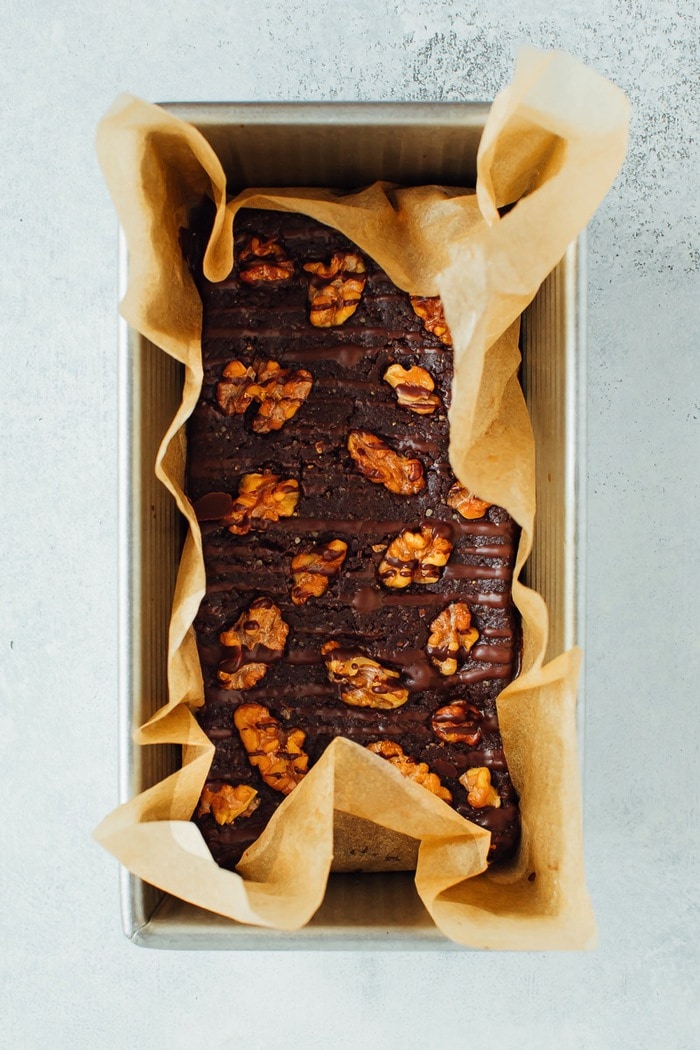 No bake brownies in a loaf pan with parchment. Brownies are topped with walnuts and a chocolate drizzle. 