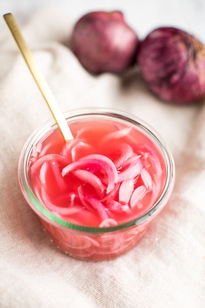 Pickled red onions in a weck jar with a gold fork and full red onions in the background.