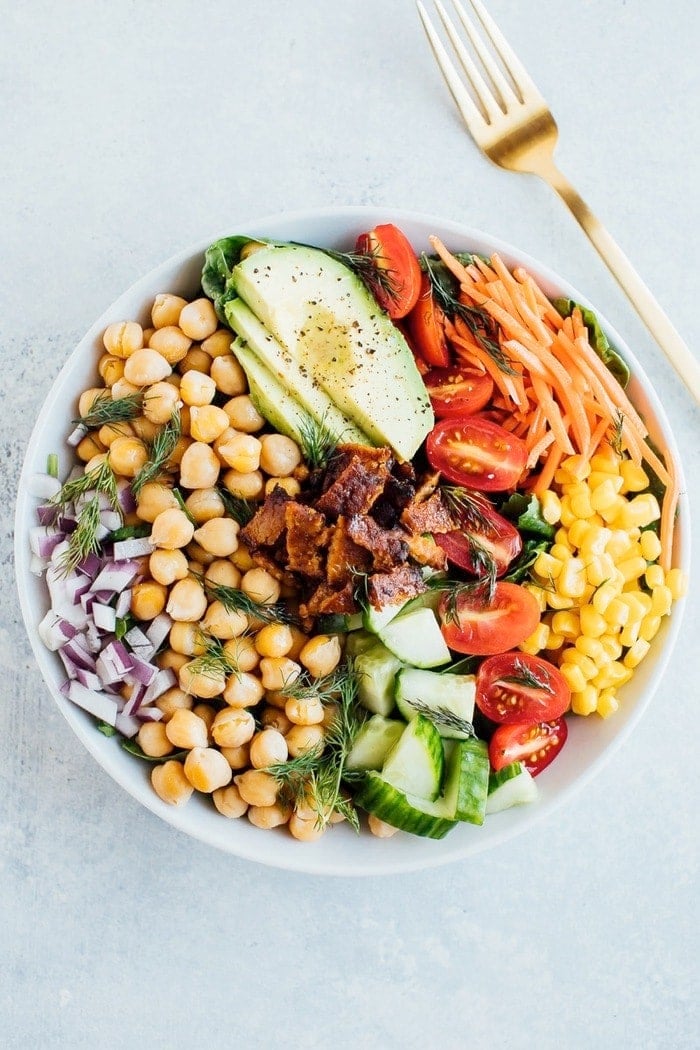 Vegan cobb salad with tempeh bacon and chickpeas in a white bowl with a gold fork. 