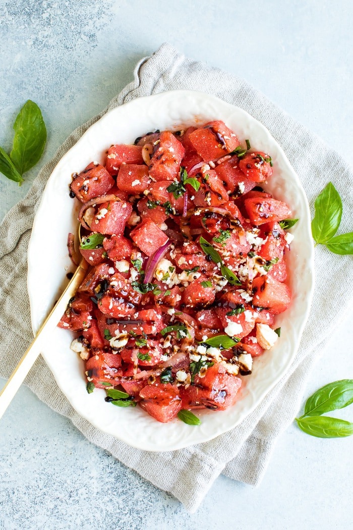 Watermelon Feta Salad with Balsamic Reduction in a white serving dish on top of a natural tan napkin. Fresh basil leaves scattered around the dish for decoration. 