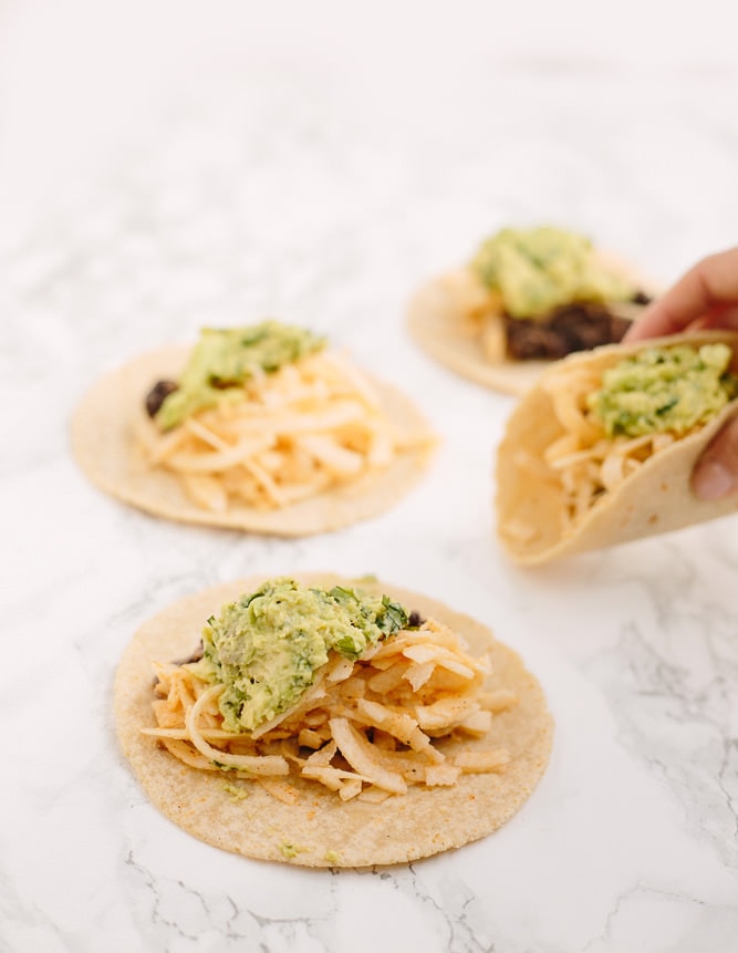 Marble counter top with 4 tacos. Hand is picking up a taco. The tacos are soft tacos with black beans, jicama, and avocado mash.