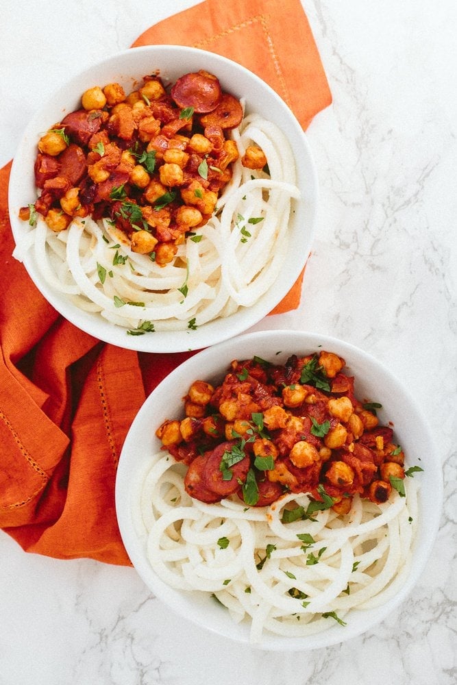 Chickpeas and chorizo in a bowl on top of jicama spiralized noodles. Two bowls of food are on top of a cloth napkin.