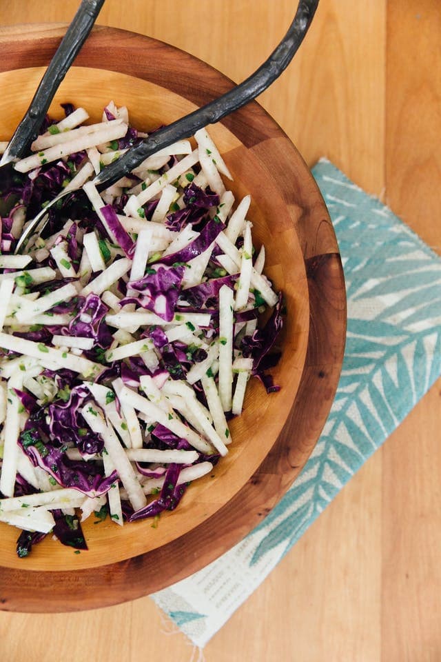 Wooden bowl filled with jicama and purple cabbage slaw with two metal serving spoons. 