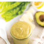 Glass jar filled with healthy avocado caesar dressing. Jar on a table next to a spoon, lettuce and an avocado.