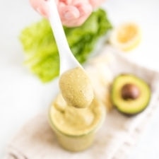 Hand holding a spoon full of avocado caesar dressing above a jar with the dressing on a table with lettuce and an avocado.