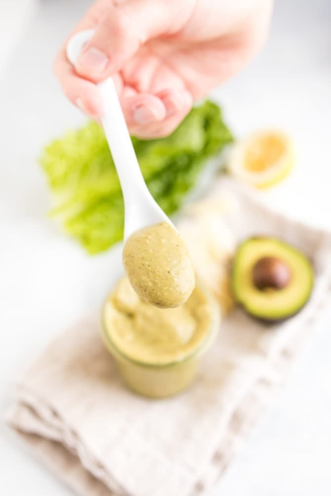 Hand holding a spoon full of avocado caesar dressing above a jar with the dressing on a table with lettuce and an avocado.