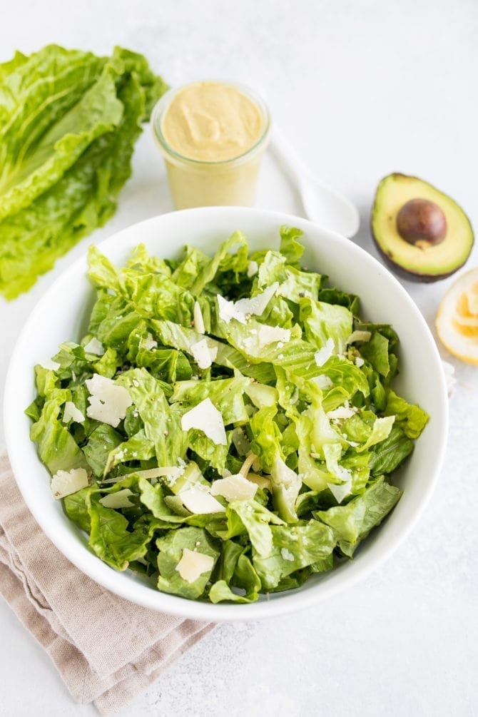 Bowl filled with romaine lettuce. Next to the bowl is a head of lettuce, a jar of avocado caesar dressing, and a half avocado.