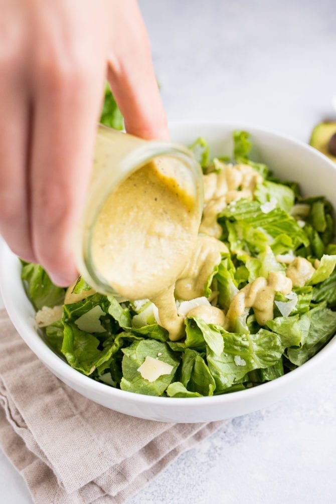 Hand pouring a jar of avocado caesar dressing over a bowl of romaine lettuce with parmesan cheese.