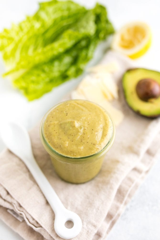 Glass jar filled with avocado caesar dressing on a table with a spoon, napkin, lettuce and avocado.