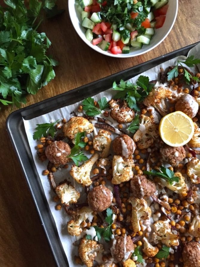 Sheet pan lined with parchment paper with baked middle eastern turkey meatballs, chickpeas, cauliflower and parsley. Sheet pan is on a wooden table with a bunch of parsley and a bowl of cucumber tomato salad.