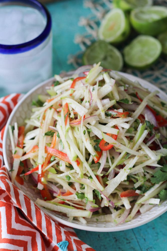 Jicama and apple slaw in a bowl next to a water glass and a bunch of limes.