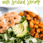 Kale caesar salad topped with grilled shrimp, avocado, sweet potatoes and parmesan cheese shavings in a white bowl. Text on top fo the image says "Kale Caesar Salad with Grilled Shrimp"