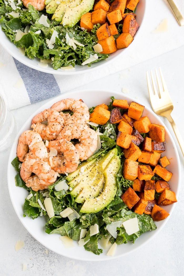 Kale caesar salad topped with grilled shrimp, avocado, sweet potatoes and parmesan cheese shavings in a white bowl with a gold fork.