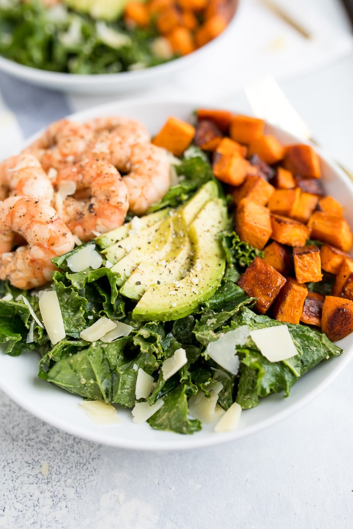 Kale caesar salad topped with grilled shrimp, avocado, sweet potatoes and parmesan cheese shavings in a white bowl.