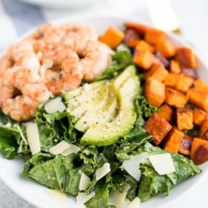 Kale caesar salad topped with grilled shrimp, avocado, sweet potatoes and parmesan cheese shavings in a white bowl.