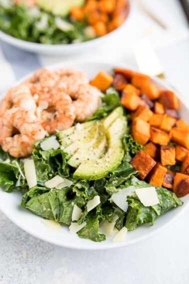 Kale caesar salad topped with grilled shrimp, avocado, sweet potatoes and parmesan cheese shavings in a white bowl.