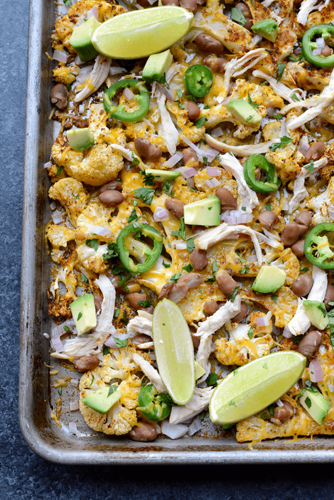 Sheet pan with baked cauliflower nachos topped with cheese, pinto beans, jalapeño slices, avocado chunks, shredded chicken, red onion, and lime slices.