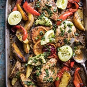 Sheet pan with baked chicken, peppers, potatoes, avocado slides and lime halves.