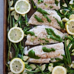 Sheet pan with baked cod, green beans, zucchini, lemon slices and an herb lemon sauce.