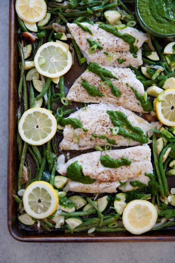 Sheet pan with baked cod, green beans, zucchini, lemon slices and an herb lemon sauce.