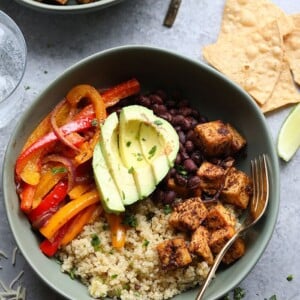 Bowl with peppers, avocado, black beans, baked Mexican tofu, quinoa. Silver fork in the bowl. The table has tortilla chips.