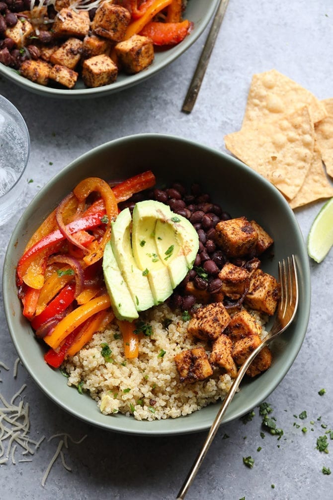 Bowl with peppers, avocado, black beans, baked Mexican tofu, quinoa. Silver fork in the bowl. The table has tortilla chips.