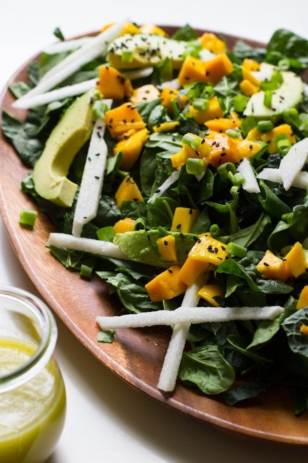 Wood serving plate with a spinach, avocado, mango and jicama salad sprinkled with sesame seeds. A glass jar of lime salad dressing is on the side.