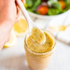 Hand holding spoon with lemon dressing over a jar of lemon dressing. Salad in the background.
