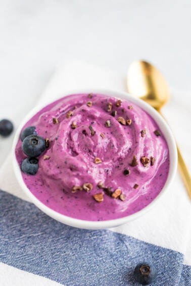 Bowl of blueberry and almond butter frozen yogurt toped with blueberries and cocao nibs. Gold spoon on the table.