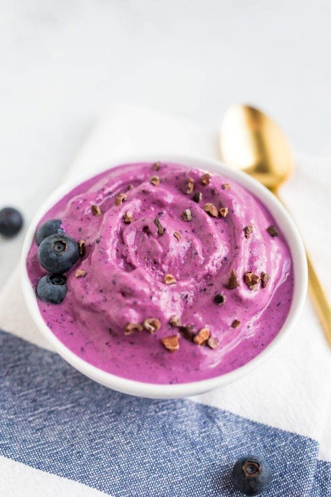 PIN THIS --> 3-Ingredient Blueberry Frozen Yogurt Bowl of blueberry and almond butter frozen yogurt toped with blueberries and cocao nibs. Gold spoon on the table.