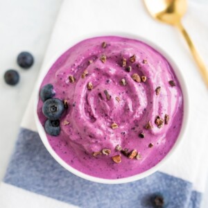 Bowl of blueberry and almond butter frozen yogurt toped with blueberries and cocao nibs. Gold spoon on the table.